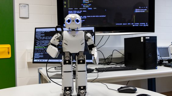 Ein Roboter steht auf einem Tisch, im Hintergrund sind Bildschirme mit Code zu sehen.