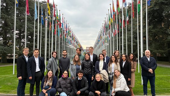 Eine Gruppe von ca. 20 Personen steht vor den Flaggen der UN-Staaten. Die Abgebildeten lächeln in die Kamera