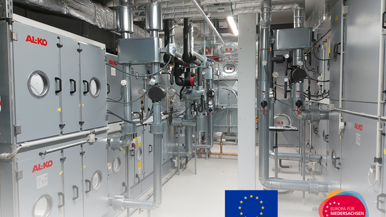 Das Bild zeigt eine moderne raumlufttechnische Anlage (RLT-Anlage) in einem Technikraum. Mehrere silberne Lüftungskanäle verlaufen über kompakten technischen Geräten mit Anzeigeelementen. Im Vordergrund sind sauber verlegte Rohre und Kabel zu sehen. Die Aufnahme ist hell und vermittelt den Eindruck moderner Gebäudetechnik.