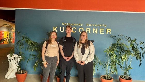 Drei Studierende stehen vor einer blauen Wand auf der in goldenen Buchstaben “Kathmandu University KU CORNER” steht. Die drei stehen vor sechs großen grünen Topfpflanzen und blicken lächelnd in die Kamera.