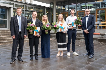 Gruppenbild mit den vier Preisträgerinnen und Preisträgern sowie Dekan und Studiendekan. Die Abgebildeten lächeln in die Kamera. Die Ausgezeichneten halten ihre Awards und Blumensträuße.